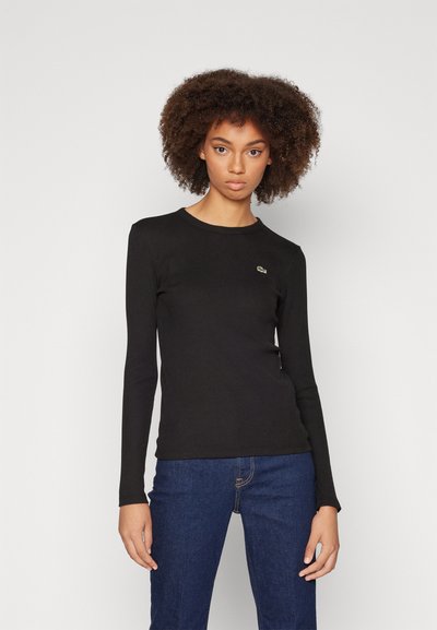 T-shirt noir à manches longues avec texture côtelée, col rond et petit logo vert sur la poitrine, associé à un jeans en denim bleu.