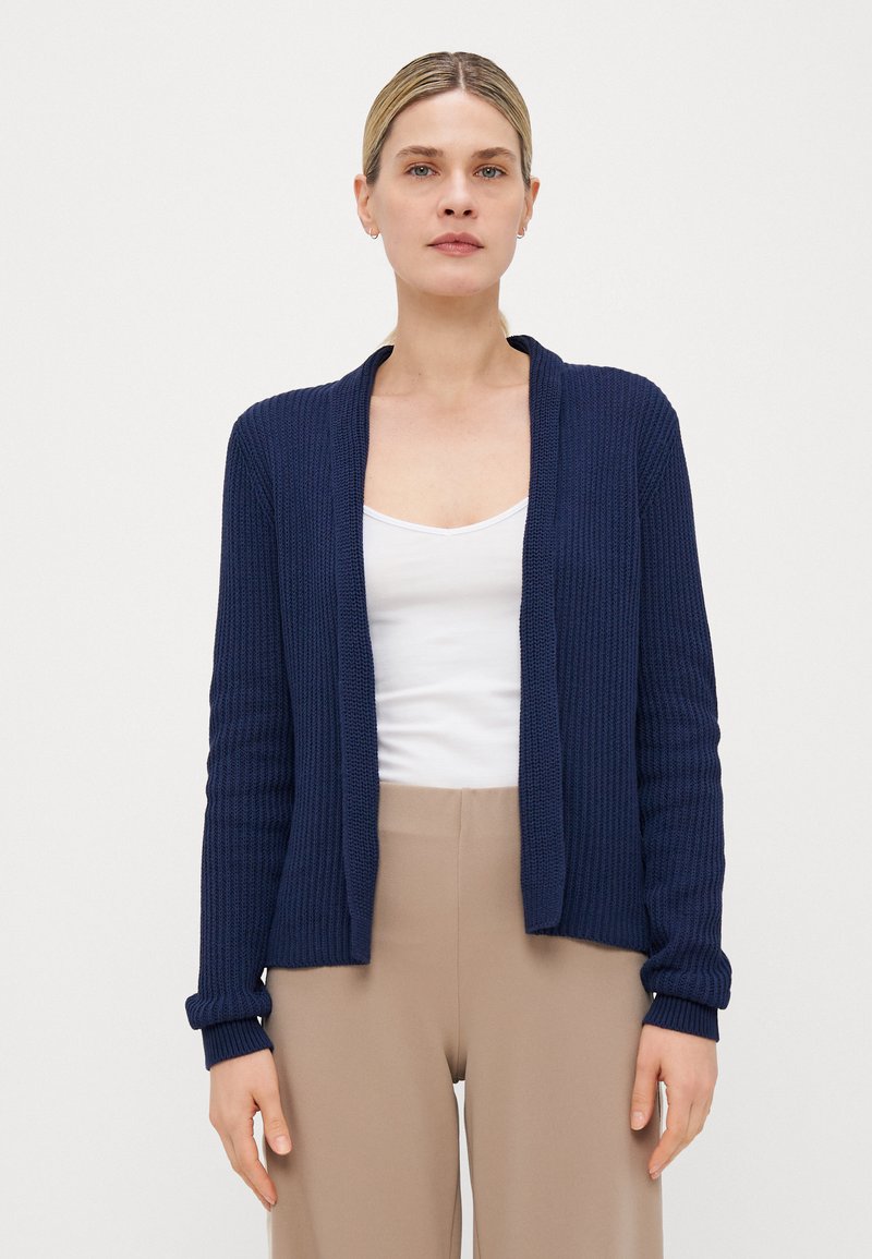 Marineblå strikket cardigan med åpen front, ribbet tekstur, lange ermer og mansjettdetaljer, båret over en hvit topp og beige bukser.