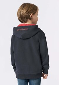 Bambino con capelli corti castani che indossa un hoodie nero con bordo rosso e il testo "U.S. POLO ASSN." sulla parte posteriore del cappuccio, di spalle.