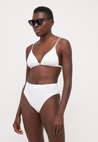 Bílé bikiny s trojúhelníkovým vrškem na tenké ramínka a vysokými kalhotkami. Hladká tkanina s minimalistickým designem a malou logovou detailní úpravou.
