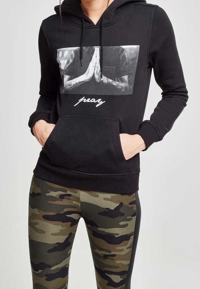 Personne portant un sweat à capuche noir avec un motif de mains en prière et le texte "pray", associé à des leggings camouflage verts et marron. Mains dans les poches du sweat.