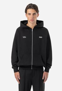 Crna zip-up majica s kapuljačom napravljena od mješavine pamuka, s prednjim džepovima i izveznim logotipima na prsima. Uključeni su rebrasti manžeti i rub.