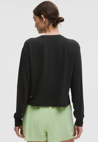 Čierny dlhý rukáv s cropped dizajnom, hladkou textúrou a voľným strihom, v kombinácii s svetlozelenými šortkami. Obsahuje logo na leme.