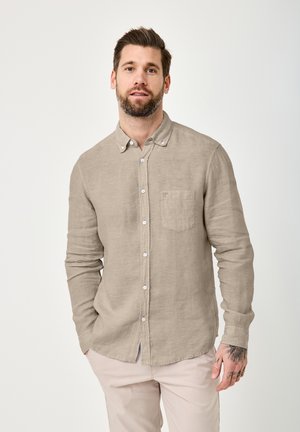 Man with beard wearing beige button-up shirt and light-colored pants, standing with one hand in pocket against plain background.