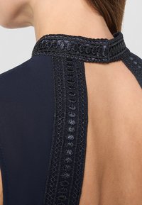 Gros plan sur le cou et le haut du dos d'une femme portant un vêtement bleu marine avec un dos en forme de trou de serrure et un col orné de dentelle noire brillante et complexe.