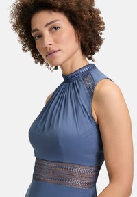 Ärmelloses Kleid mit hohem Kragen aus blauer Stoff; verfügt über geraffte Falten und Spitzenakzente am Hals- und Taillenbereich, glatte Textur.