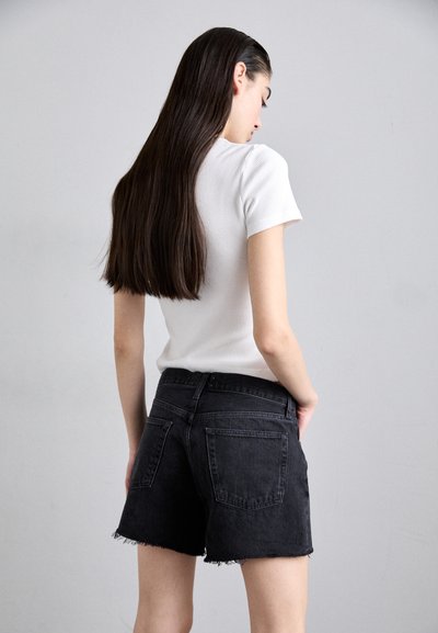 Žena s dlhými tmavými vlasmi otočená bokom, oblečená v bielej krátkoročkej košeli a čiernych rifľových šortkách s ošúchanými lemami.