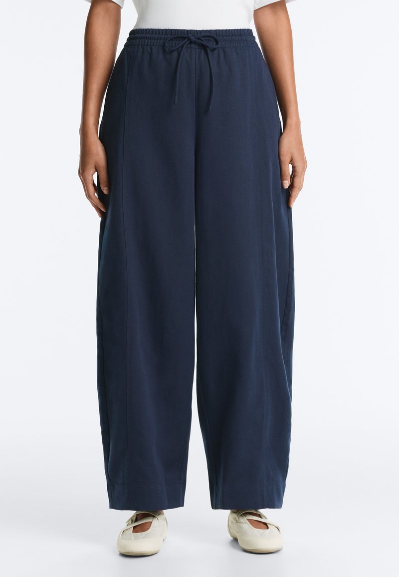 Pantalon ample bleu marine avec taille élastique et cordon, porté avec un haut blanc et des chaussures crème à enfiler.
