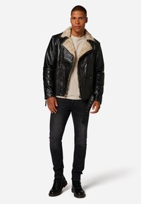 Svart läderjacka med en krämfärgad fårskinnskrage, dragkedja fram och sidofickor, bärs över en ljusbeige skjorta och mörka jeans.