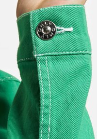 Primer plano de un puño de mezclilla verde con costuras blancas y botón metálico etiquetado "ONLY" que sujeta un lazo blanco.