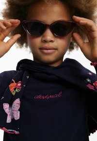 Schwarze cat-eye Sonnenbrille, marineblaues geripptes Shirt mit rosa gesticktem "desigual" und einem mehrfarbigen blumenmusternden Schal.