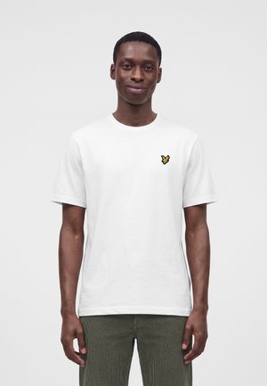 Homme portant un t-shirt blanc uni à manches courtes avec un petit logo jaune d'oiseau sur la poitrine gauche et un pantalon vert olive.