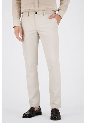 Mann trägt beige, schmal geschnittene Hose, hellbeiges Hemd und schwarze Leder-Loafer, steht mit einer Hand in der Tasche.