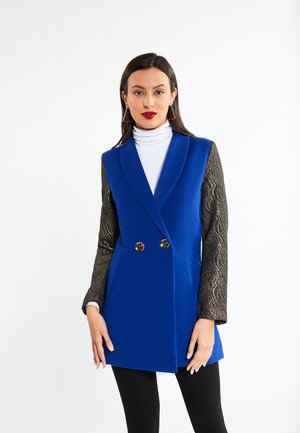 Nainen, jolla on sininen kaksirivinen takki, jossa on teksturoidut mustat hihat, valkoinen poolopaita, mustat housut ja kolmionmuotoiset kultaiset korvakorut, seisoo paikallaan.