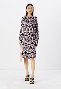 Schwarzes und pinkes blumenbedrucktes Kleid, lange Ärmel, weicher Stoff, lockere Passform, bis zur Mitte des Oberschenkels reichend. Das Model trägt eine hellfarbige Handtasche.