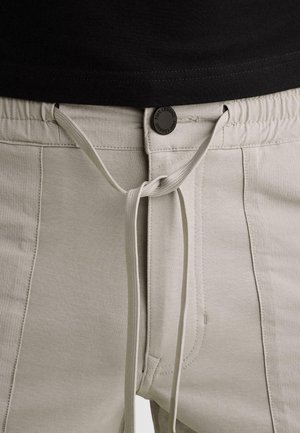 Pantalon beige clair avec ceinture élastique, bouton avant étiqueté « PME Legend » et lien noué au-dessus de l'ourlet de la chemise noire.