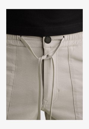 Pantalon beige clair avec ceinture élastique, bouton avant étiqueté « PME Legend » et lien noué au-dessus de l'ourlet de la chemise noire.