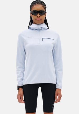 Vrouw die een lichtblauwe lange mouw pullover met ritssluiting en zwarte fietsshorts draagt, met gevlochten haar en zwarte sportzonnebril.
