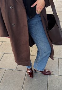 Brūns vilnas mētelis ar lieliem pogām, zilas vidēja augstuma džinsus un bordo kvadrātveida pirkstai apavus. Ir klāt brūna rokassoma. Zemē ir teksturēts bruģis.