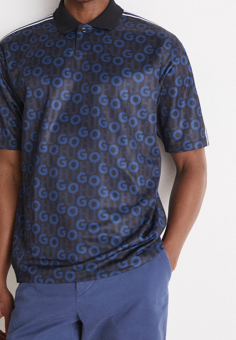 Homme portant un polo foncé avec un motif texte bleu "GO" sur toute la surface et un pantalon bleu, debout avec une main dans la poche.