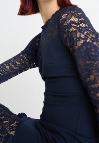 Marinblå klänning med långa spetsärmar med blommönster, en figursydd silhuett och en scalloped halsringning. Slät tygtextur genom hela plagget.