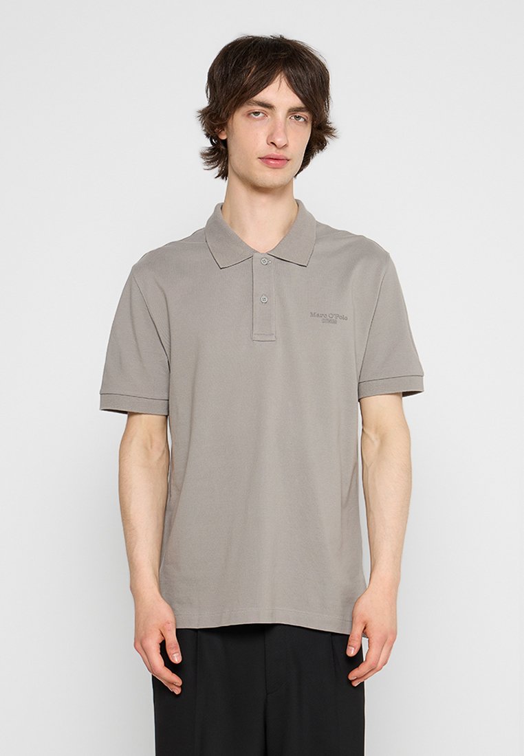 Junger Mann in einem taubfarbenen kurzärmeligen Poloshirt und schwarzen Hosen, der vor einem schlichten hellen Hintergrund steht.