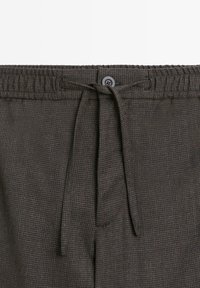 Pantaloni a scacchi marroni con vita elastica, cordoncino e chiusura a bottone. Il tessuto ha una leggera texture.