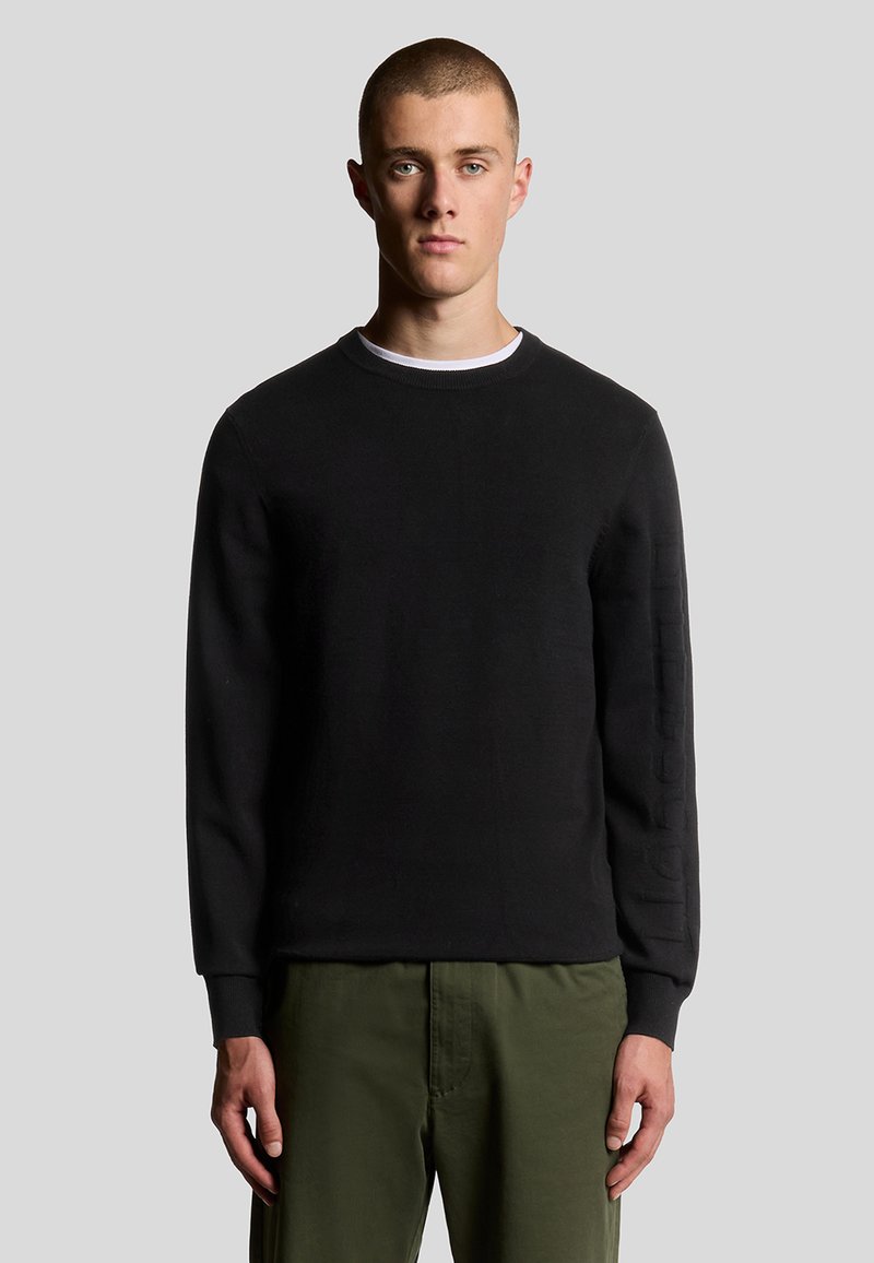 Zwarte sweatshirt met ronde halslijn, lange mouwen en textuuraccenten op de mouwen. Gecombineerd met olijfgroene broek.