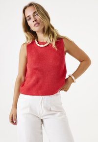 Roter, gerippter ärmelloser Pullover mit hohem Kragen, kombiniert mit weißen, plissierten Hosen. Ergänzt durch eine grobe, neutrale Halskette.