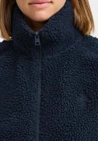 Mörk marinblå fleecejacka med hög krage, hel dragkedja, texturerat tyg och en liten logotyp på vänster sida. Mjuk, varm look.