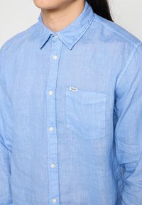 Camicia di lino azzurro chiaro con colletto classico, bottoni sul davanti, taschino singolo sul petto e piccolo etichetta del marchio sulla tasca. Texture morbida.