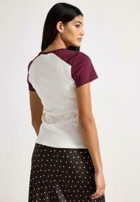 T-shirt med korta ärmar, vit kropp med vinröda raglanärmar, gjord av mjuk bomull, kombinerad med en prickig brun kjol.