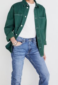 Person som bär en grön skjorta över en vit t-shirt, i kombination med blå jeans, en hand i fickan, vit bakgrund.