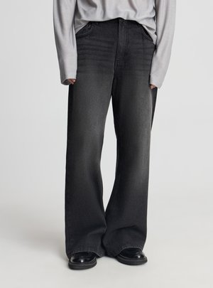 Persona che indossa jeans neri ampi e scarpe nere, con le mani parzialmente coperte da una maglia a maniche lunghe grigio chiaro, in piedi su sfondo uniforme.