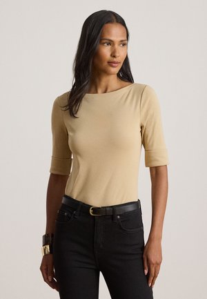 Kvinne med langt mørkt hår iført en beige topp med trekvart ermer stukket ned i svarte jeans med svart belte og gullspenne.