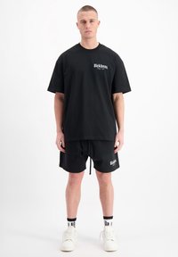 Zwarte katoenen T-shirt met korte mouwen en logo-opdruk op de borst, gecombineerd met zwarte shorts met een trekkoord en logo-detail. Witte sneakers.