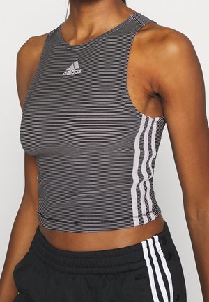 Frau trägt ein schwarz-weiß gestreiftes ärmelloses Adidas Crop-Top und schwarze Hose mit weißen Adidas-Streifen.