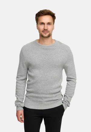 Grå stickad tröja med ett texturerat diagonalt mönster, rundad halsringning och ribbade manchetter. Kombinerad med svarta byxor, vilket ger en enkel ensemble.
