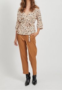 Blouse à fleurs beige avec des manches courtes et une ceinture à nouer, associée avec un pantalon marron fuselé et des bottines noires.