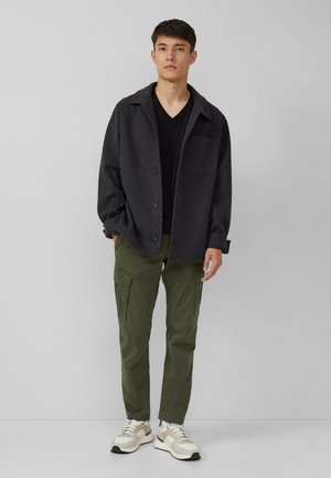 Dunkelgraue Button-up-Jacke über einem schwarzen V-Ausschnitt-T-Shirt, kombiniert mit grünen Cargo-Hosen und grau-weißen Sneakers. Details des lässigen Outfits sind sichtbar.