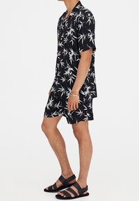 Camisa negra de manga corta con estampado de bambú blanco, con cierre de botones, combinada con pantalones cortos a juego y sandalias negras.