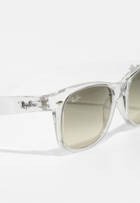 Ochelari de soare din plastic transparent cu lentile gri, formă rectangulară și o branding subtil Ray-Ban pe ramă și marginile lentilelor.