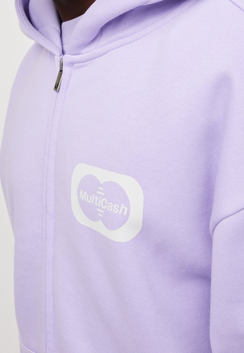 Gros plan d'une personne portant un sweat à capuche zippé violet clair avec un logo blanc "MultiCash" sur la poitrine.