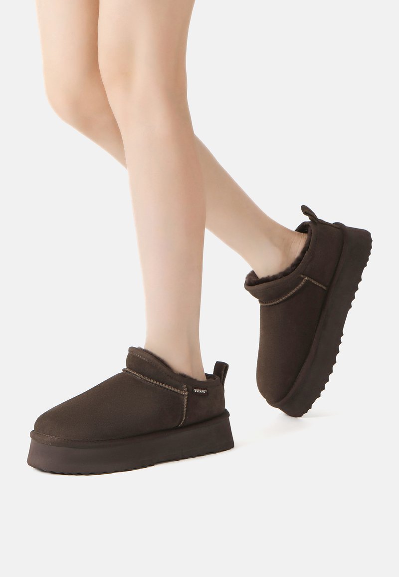 Füße, die dunkelbraune, gefütterte Schlupfschuhe mit dicken Sohlen tragen, vor einem schlichten hellen Hintergrund.