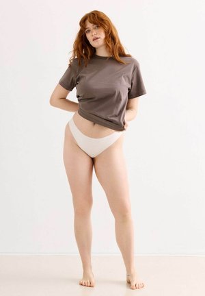 Rothaarige Frau steht barfuß, trägt ein braunes T-Shirt und helle, hochgeschnittene Unterwäsche vor einem einfarbigen weißen Hintergrund.