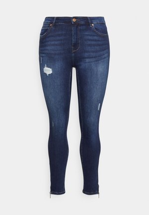 Donkere blauwe skinny jeans met licht versleten stukjes, voorzakken, knoopsluiting en enkelritsen op een effen achtergrond.