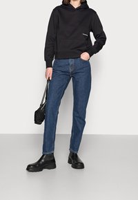 Sudadera con capucha negra y bolsillo frontal, combinada con jeans rectos azules y botas negras de tobillo. Diseño simple con un aspecto casual.