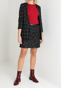 Blazer de cuadros negros sobre una camiseta de canalé roja, combinado con una falda negra con estampado y botines burdeos brillantes.