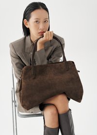 Mujer sentada en una silla de metal, vestida con blazer gris y botas negras, sosteniendo un gran bolso de ante marrón con asas dobles.