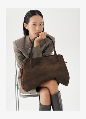 Woman seated on metal chair wearing gray blazer and black boots, holding large brown suede handbag with double handles.